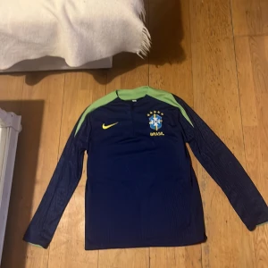 Blå långärmad Brasilien fotbollströja från Nike - Säljer en blå långärmad fotbollströja från Nike med Brasilien-emblem på bröstet. Tröjan har gröna detaljer vid kragen och ärmsluten samt en gul Nike-logga. Perfekt för fotbollsfans!
