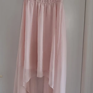 Ljusrosa långklänning, Gina tricot - Elegant ljusrosa långklänning med volangdetaljer längs axelbanden och en vacker öppen rygg. Klänningen har en luftig passform och faller fint med ett asymmetriskt snitt. Perfekt för speciella tillfällen. Storlek 34 men passar 36