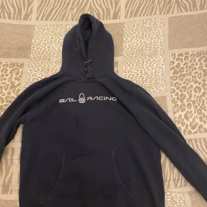 Mörkblå hoodie från Sail Racing - Säljer en snygg mörkblå hoodie från Sail Racing. Den har en stor logga på bröstet och en praktisk ficka framtill. Perfekt för kyliga dagar och avslappnade stunder. Huvan har dragsko för justering. Passar både till vardags och för sportiga aktiviteter.Det finns också lite fläckar på den.