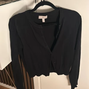 Svart kofta från H&M - Säljer en klassisk svart kofta från H&M i bra skick. Den har långa ärmar och knappar framtill. Passar till allt och är superbekväm!