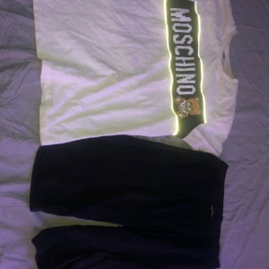 Vit t-shirt och shorts från Moschino Teen - Säljer en cool vit t-shirt från Moschino Teen med ett stort Moschino-tryck och en söt nalle på framsidan. Perfekt för en avslappnad stil och passar bra till både jeans och shorts. T-shirten är i bra skick och redo för nya äventyr!