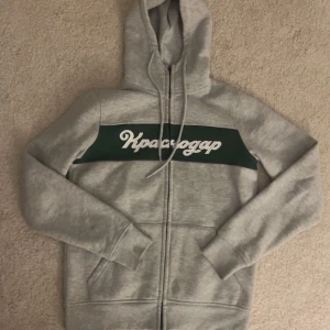 Grå hoodie med grön detalj - Säljer en grå zip med en grön rand och vit text på bröstet. Den har en svart dragkedja framtill och en mysig luva🩷Skriv vid funderingar och fler bilder🩷