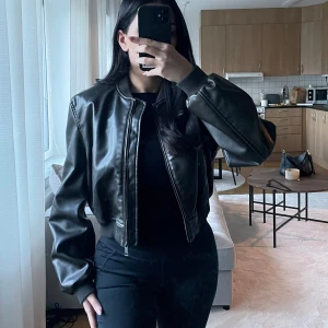 Svart skinnjacka - Säljer en snygg svart skinnjacka i mycket bra skick. Den har en klassisk bomberjacka-stil med dragkedja framtill och ribbade muddar vid ärmslut och nederkant. Perfekt för höst och vår, och ger en cool look till vilken outfit som helst. Passar bra till både vardag och fest!