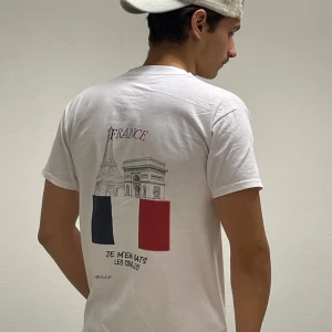 Vit t-shirt med franskt motiv - Säljer en vit t-shirt med tryck av Eiffeltornet och Triumfbågen samt franska flaggan. Texten 'Je m'en bats les couilles' ger en kaxig touch. Perfekt för dig som vill ha en casual och unik stil. Passar bra till jeans eller shorts! tröjan är i storlek M och modellen är 190 och 77kg