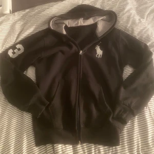 Svart hoodie från Ralph Lauren - Säljer en svart hoodie från Ralph Lauren med dragkedja och huva. Den har en vit logga på bröstet och siffran 3 på ärmen. Perfekt för en casual look eller mysiga dagar hemma. Passar bra till både jeans och mjukisbyxor.