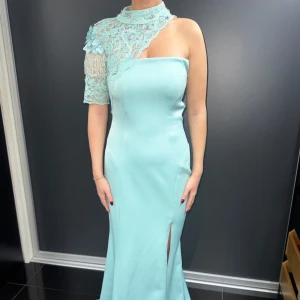 Mintgrön långklänning med spetsdetaljer - Elegant mintgrön långklänning med en unik one-shoulder design och spetsdetaljer. Klänningen har en slits framtill och en vacker öppen rygg. Perfekt för festliga tillfällen!