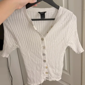 Vit ribbad topp från Lindex - Säljer en superfin vit ribbad topp från Lindex i storlek XS. Den har korta ärmar och knappar framtill, perfekt för vår och sommar. Toppen är i mycket bra skick och passar både till vardags och fest. 