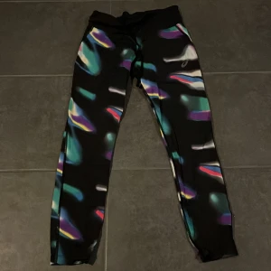 Mönstrade träningsbyxor från Asics - Säljer ett par coola träningsbyxor från Asics i svart med färgglatt abstrakt mönster. De har en bekväm passform och är perfekta för träning. Byxorna har en elastisk midja med snörning för bästa passform. Perfekta för att sticka ut på gymmet!