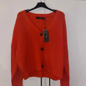 Orange stickad kofta från Vero Moda - Säljer en superfin orange stickad kofta från Vero Moda. Den har en v-ringad design med stora knappar framtill. Perfekt för höst och vinter, och den är i nytt skick, inte använd!! Koftan är både mysig och stilren, perfekt för att hålla värmen med stil!