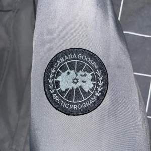 Säljer en snygg grå jacka från Canada Goose, perfekt för kyliga dagar. Jackan är i mycket bra skick och har en klassisk design med broderad logga på ärmen. Tillverkad i Kanada med högkvalitativa material. Passar perfekt för både höst och vinter.