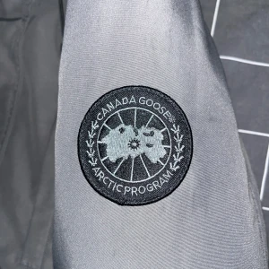 Grå jacka från Canada Goose - Säljer en snygg grå jacka från Canada Goose, perfekt för kyliga dagar. Jackan är i mycket bra skick och har en klassisk design med broderad logga på ärmen. Tillverkad i Kanada med högkvalitativa material. Passar perfekt för både höst och vinter.