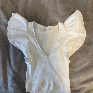 Vit ribbad topp från Zara - Säljer en superfin vit ribbad topp från Zara i storlek XS. Den har en djup v-ringning och volangdetaljer på ärmarna som ger en romantisk touch. Perfekt för sommardagar eller en utekväll. Toppen är i mycket bra skick och redo för nya äventyr! 🤍