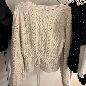 Beige stickad tröja från Zara - Säljer en superfin beige stickad tröja från Zara. Den har ett snyggt flätmönster och en knytning i midjan som ger en fin passform. Perfekt för höst och vinter! Tröjan är i mycket bra skick 