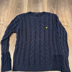 Mörkblå stickad tröja från Lyle & Scott - Säljer en snygg mörkblå stickad tröja från Lyle & Scott med kabelstickat mönster. Den har en gul logga på bröstet och långa ärmar. Perfekt för kyliga dagar och ger en stilren look. Passar både till vardags och lite finare tillfällen.