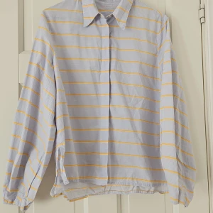 Randig skjorta med gula och vita ränder - Säljer en superfin randig skjorta med gula och vita ränder. Skjortan har långa ärmar och en klassisk krage. Perfekt för både vardag och lite mer uppklädda tillfällen. Den är i bra skick och redo för en ny ägare!