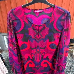 Färgglad mönstrad blus från Indiska - Säljer en superfin och färgglad klänning/tunika från Indiska i storlek XL. Blusen har ett iögonfallande mönster i rosa, lila och svart. Den är långärmad och perfekt för både vardag och fest.
