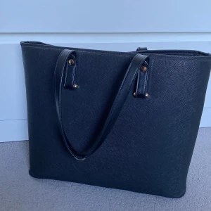 Svart axelväska i syntetmaterial - Säljer en stilren svart axelväska i syntetmaterial. Väskan har en enkel och elegant design med två handtag och en dragkedja upptill. Perfekt för både vardag och jobb!