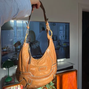 Brun axelväska i skinn - Säljer en snygg brun axelväska i skinn med coola detaljer. Väskan har en rund form och är perfekt för vardagsbruk. Den har en dragkedja upptill och dekorativa sömmar som ger den en unik look. Perfekt för höst💓🤩