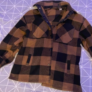 Rutig overshirt i brun och svart - Säljer en oversize jacka i brun och svart med rutigt mönster. Den har fyra fickor  och knappar framtill. Perfekt för höst och vår. Använd ett fåtal gånger. 