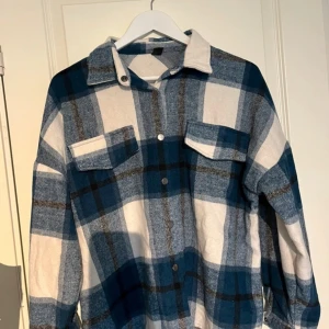 Blå och vit rutig overshirt - Säljer en snygg blå och vit rutig overshirt med långa ärmar. Den har knappar framtill och två bröstfickor som ger en cool look. Perfekt för höst och vår när du vill ha något stilrent och bekvämt. Passar både till vardags och lite mer avslappnade tillfällen.