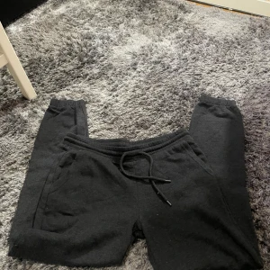 Svarta mjukisbyxor från H&M Divided - Säljer ett par svarta mjukisbyxor från H&M Divided i storlek S. Byxorna är gjorda av en mjuk bomulls- och polyesterblandning, perfekt för en avslappnad dag hemma eller på gymmet. De har en bekväm resårmidja med snörning för justerbar passform.