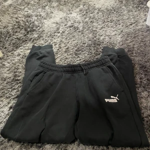 Svarta mjukisbyxor från Puma - Säljer ett par svarta mjukisbyxor från Puma i storlek M. Byxorna har en elastisk midja och är superbekväma för chill eller träning. De har fickor på sidorna och Puma-loggan tryckt på ena benet. Perfekta för en avslappnad stil!