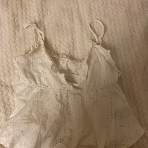 Vit ärmlös topp med spetsdetaljer - Säljer en söt vit topp med spetsdetaljer och volanger. Den är ärmlös och har en luftig passform, perfekt för sommardagar. Toppen har ett fint hålmönster och knytband vid axlarna.