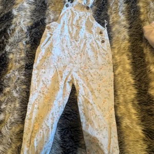 Overall Newbie - Säljer en superfin blommig byxdress från Newbie. Den är ljusblå med ett sött blommönster i rosa och grönt. Byxdressen har justerbara axelband och knappar på sidorna för enkel påklädning. Perfekt för vår och sommar!