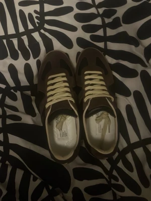 Maison Margiela Gats Bruna - Säljer ett par Maidon Marhiela gats i brunt med beigea snören och detaljer. Skorna har en låg profil och är gjorda i en mix av mocka och läder. Perfekta för dig som gillar en klassisk och clean look.