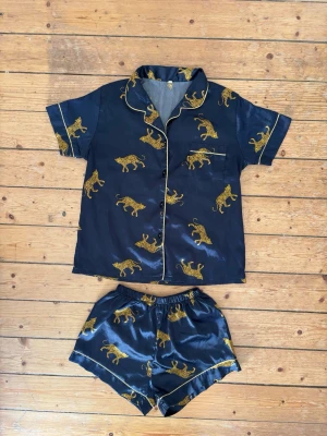 Mörkblå satinpyjamas med leopardmönster storlek S - Snygg tvådelad pyjamas i mörkblå satin med kortärmad skjorta och shorts. Setet har ett gult leopardmönster och tunna gula passpoaler längs kanterna. Skjortan har knappar framtill och krage, shortsen har resår i midjan. Perfekt skick! Storlek S