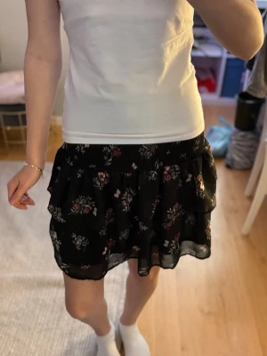 Svart blommig volangkjol - Säljer en svart kortkjol med blommigt mönster och volangdetaljer. Kjolen har en luftig och flowy passform, perfekt för varmare dagar. Resår i midjan för extra komfort och snygg passform.