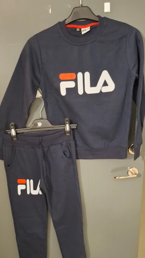 Mörkblåa mjukisbyxor från FILA - Mörkblåa mjukisbyxor från FILA med resår i midjan och fickor på sidorna. Byxorna har en stor vit och orange FILA-logga på ena benet. Tillverkade i mjukt bomullsmaterial för maximal komfort och avslappnad stil.