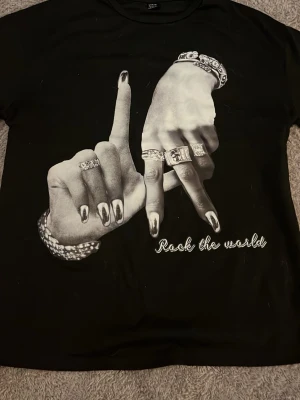 Svart t-shirt med tryck och text - Cool svart t-shirt med stort tryck av två händer med smycken och texten 'Rock the world' framtill. T-shirten har korta ärmar och en klassisk passform. Perfekt för dig som vill sticka ut med en unik och edgy stil.