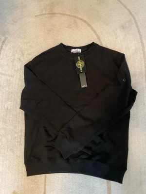 Svart stickad tröja från Stone Island - Snygg svart stickad tröja från Stone Island med klassisk rund halsringning och ribbade muddar. Tröjan har en diskret logotyp på ärmen och en ikonisk Stone Island patch medföljer. Perfekt för dig som gillar stilrena och tidlösa plagg.