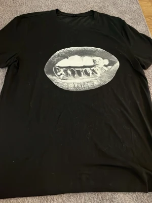 Svart t-shirt med läppar-tryck - Säljer en svart t-shirt med ett stort, coolt tryck av läppar och tänder i vitt på bröstet. T-shirten har klassisk passform och korta ärmar. Perfekt för dig som vill sticka ut med en unik design.