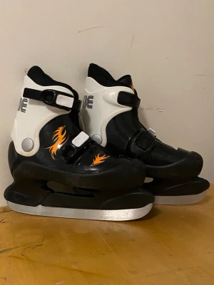 Svarta och vita skridskor med spännen - Säljer ett par svarta och vita skridskor med coola orange flames på sidorna. Skridskorna har två justerbara spännen för bra passform och robust plastskal. Perfekta för dig som vill sticka ut på isen!