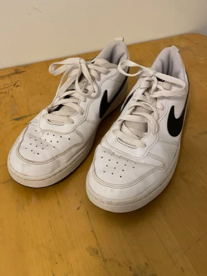 Vita Nike sneakers med svart swoosh - Säljer ett par klassiska vita Nike sneakers med svart swoosh på sidan. Skorna har lågt skaft, vita snören och ventilerande hål på tån. Yttermaterialet är slätt och de har en bekväm, vadderad insida. Perfekta för en clean och sportig stil.