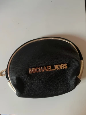 Svart rund liten väska Michael Kors - Liten rund väska från Michael Kors i svart med guldfärgade detaljer och dragkedja. Väskan har en tydlig metallogo framtill och guldfärgad kant runtom. Perfekt för småprylar eller som accessoar.