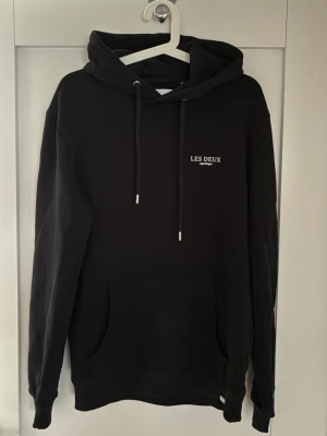 Hoodie från Les Deux - Oanvänd hoodie från Les Deux med vit logga på bröstet och dragsnören med metalldetaljer. En kvalitetshoodie, perfekt för en avslappnad stil. 
