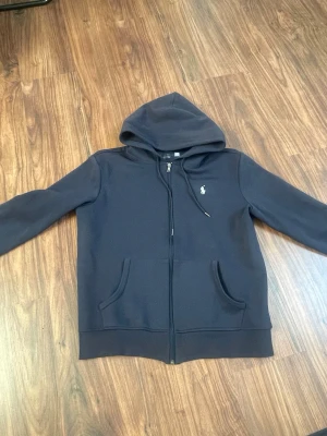 Mörkblå Polo Ralph Lauren set - Mörkblått set från Polo Ralph Lauren med dragkedja, huva och två fickor framtill. Klassisk vit logga broderad på bröstet. Perfekt för en avslappnad och sportig stil. Tillverkad i mjukt bomullsmaterial.