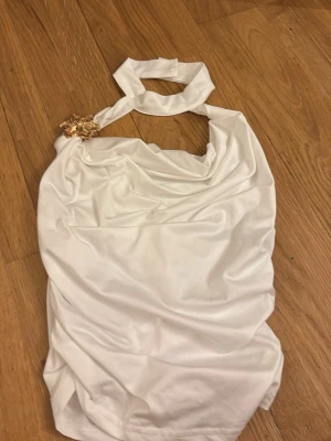 Vit halterneck topp med blomdetalj - Säljer en vit halterneck topp i glansig polyester med en snygg guldfärgad blomdetalj på axeln. Toppen har draperad framsida och är ärmlös, perfekt för dig som vill sticka ut lite extra.