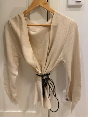 Beige omlottkofta med knytskärp - Stilren beige omlottkofta med långa ärmar och svart knytskärp i midjan. Koftan har en öppen front och är tillverkad i ett mjukt, vävt material som ger en avslappnad men trendig look. Perfekt att slänga över en topp för en snygg layering-effekt.