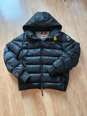 Parajumpers Sheen Svart Pufferjacka - Snygg svart pufferjacka från Parajumpers, modell Sheen. Jackan har huva, dragkedja framtill, ribbade muddar och en gul detalj vid halsen. Märkespatch på ärmen och två sidofickor. Väldigt varm och skön på kroppen. Storlek M