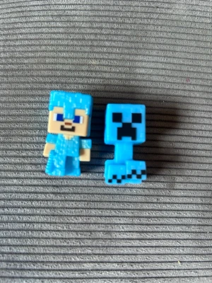 Minecraft minifigurer blå - Två små Minecraft minifigurer i plast, en föreställer en karaktär med blå rustning och den andra en blå creeper. Perfekta som samlarprylar eller dekoration till skrivbordet. Figurerna har tydliga pixlade detaljer och starka färger.