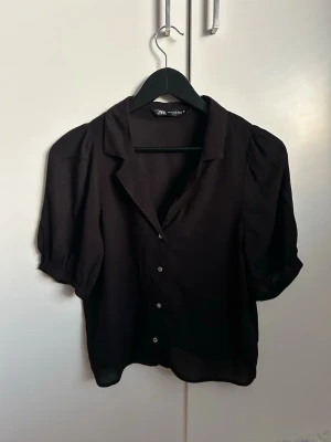 Svart blus med kort ärm från Zara - Stilren svart blus från Zara med korta puffärmar och knappar framtill. Blusen har en klassisk krage och är gjord i ett tunt, lätt och något transparent material som ger en luftig känsla.