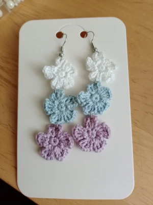 Virkat blommiga örhängen i pastell - Handgjorda örhängen med virkade blommor i vitt, ljusblått och lila. Hängande modell med tre blommor i rad, fästa på silverfärgade krokar. Perfekt accessoar för en färgglad och lekfull stil.