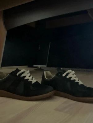 Maison margiela gats - Säljer ett par svarta sneakers med vita snören och klassisk brun gummisula. Skorna har en låg profil och är tillverkade i en mix av mocka och textil. Perfekta för dig som gillar stilrena och tidlösa sneakers.