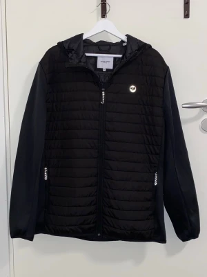 Svart pufferjacka från Jack & Jones - Säljer en svart pufferjacka från Jack & Jones med huva och dragkedja framtill. Jackan har två fickor med dragkedja och en diskret logga på bröstet. Tillverkad i lätt och värmande material, perfekt för kalla dagar. Mått: längden är ca 77cm, axelbredden är ca 51cm och ärmlängden är ca 67cm långt💕