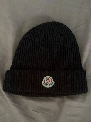 Svart ribbstickad mössa Moncler - Säljer en svart ribbstickad mössa från Moncler med uppvikt kant och klassisk Moncler-logga framtill. Mössan är gjord i ett mjukt och varmt material som passar perfekt för kyliga dagar. Enkel och stilren design som funkar till det mesta.