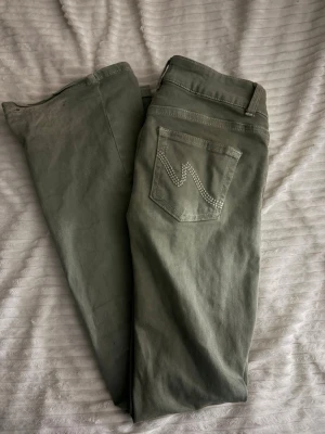 Olivgröna bootcut jeans med detaljer - Snygga olivgröna jeans med bootcut passform och dekorativa sömmar på bakfickan. Jeansen har klassisk femficksdesign och stängs med knapp och dragkedja. Perfekta för dig som vill ha en unik twist på vardagslooken.
